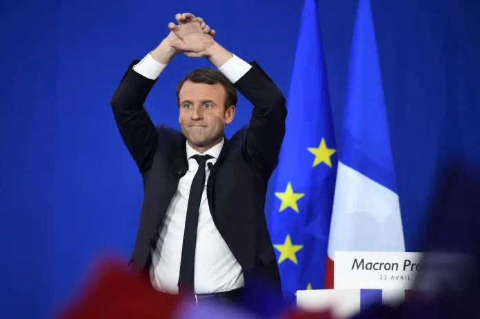1253526-emmanuel-macron-le-5-mai-2017 Emmanuel Macron sur scène en costume, les bras levés face à un drapeau français et européen